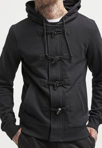 Sweat à capuche noir en matière douce, avec fermetures à bascule et poches avant. Le design comprend une poche kangourou et des détails texturés.