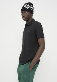 Μαύρο πόλο μπλουζάκι με μικρό λογότυπο, συνδυασμένο με πράσινο τζόγγερ. Το μοντέλο φοράει μαύρο σκουφάκι με λευκό σχέδιο. Στενή εφαρμογή.