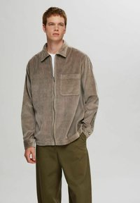 Kamjakke i taupe corduroy med lynlås foran, krave-design, lange ærmer og en enkelt brystlomme. Båret over en hvid T-shirt.