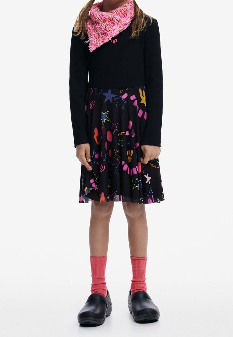 Enfant portant un haut noir à manches longues, une jupe noire avec des motifs d'étoiles colorées, une écharpe rose tricotée, des chaussettes roses et des chaussures noires, immobile.