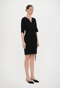 Crna haljina omotnica od mekog materijala s kratkim rukavima, V-izrezom i detaljem vezanja sa strane. Uparena s crnim cipelama na visoku petu. Jednostavan i elegantan dizajn.