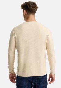 Pull à manches longues beige avec une finition texturée et un col ras du cou. Il dispose d fentes sur les côtés à l'ourlet et de poignets ajustés.