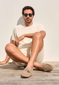 Hellbeige Mokassins mit einer Schleifenapplikation, getragen mit einem cremefarbenen kurzärmeligen Rippenshirt und passendem Short, auf einer Holzoberfläche.