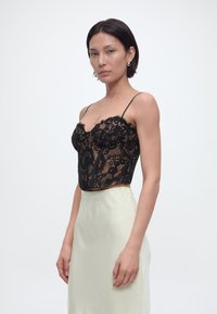 Γυναίκα με κοντά σκούρα μαλλιά, που φορά χρυσούς κρίκους στα αυτιά, μαύρο κορσέ από δαντέλα και κρεμ σατινένια φούστα, σε λευκό φόντο.