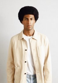 Beige katoenen jacket met knoopsluiting, twee zijzakken en een borstzak. Gedragen over een wit shirt en lichtblauwe jeans.