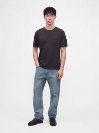 Jeune homme debout portant un t-shirt noir à manches courtes, un jean bleu clair et des chaussures noires sur un fond blanc uni.