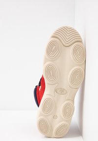 Semelle de chaussure en caoutchouc beige avec des crampons circulaires et dessus en daim rouge, positionnée verticalement contre un fond blanc d'angle.