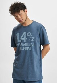 Blå bomullst-skjorte med en stor grafisk utskrift som leser "14% PREMIUM DENIM." Korte ermer, rund halslinje, avslappet passform.