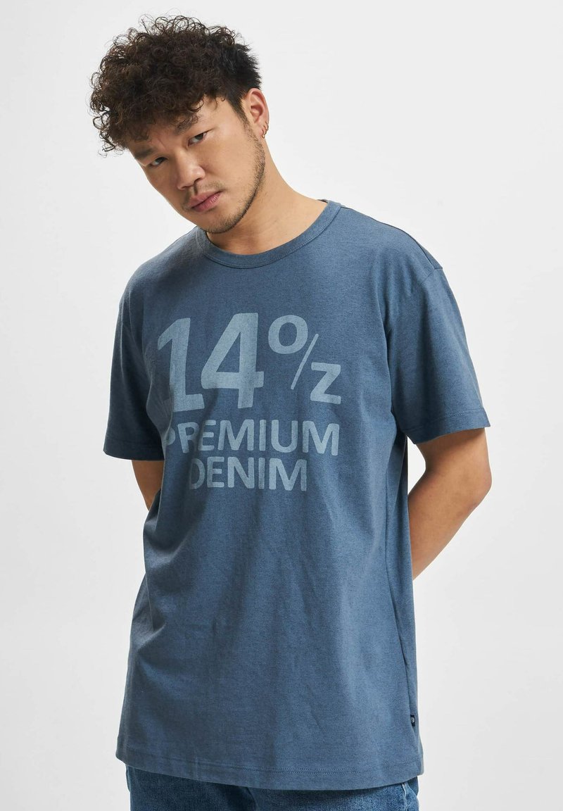 Blå bomullst-skjorte med en stor grafisk utskrift som leser "14% PREMIUM DENIM." Korte ermer, rund halslinje, avslappet passform.