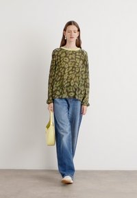 Lichtgewicht, doorschijnend groen topje met abstract patroon, ronde halslijn en geplooide mouwen; gecombineerd met blauwe wijde broek en gele tas.