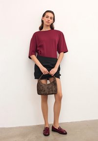 Ženska oblečena v burgundski kratkosleeved top, črno mini krilo, burgundske loafer čevlje, drži majhno rjavo torbico s potiskom ob belem zidu.