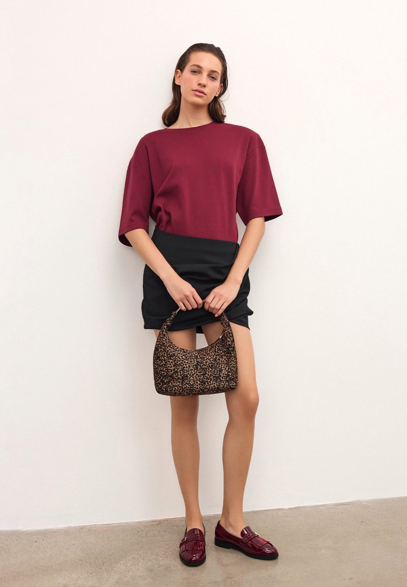 Ženska oblečena v burgundski kratkosleeved top, črno mini krilo, burgundske loafer čevlje, drži majhno rjavo torbico s potiskom ob belem zidu.