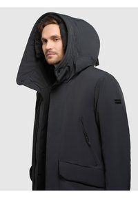 Geacă izolată neagră cu un glugă mare, structurată, închidere frontală cu fermoar, buzunare laterale și un detaliu din țesătură texturată pe mânecă.