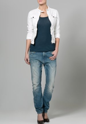 Femme debout portant une veste courte blanche, un débardeur bleu marine, un jean bleu délavé et des chaussures à talons hauts marron foncé sur fond gris.