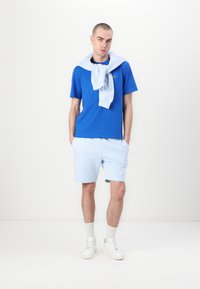 Μπλέ βαμβακερό t-shirt, ανοιχτό μπλε σορτς και ένα ανοιχτό μπλε πουλόβερ ριγμένο στον λαιμό. Το μοντέλο φοράει λευκά αθλητικά παπούτσια και κάλτσες.