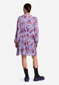 Fioletowa sukienka w kwiaty z długimi rękawami, zwężoną talią i wzorzystym materiałem zawierającym intensywne akcenty pomarańczowego, różowego i zielonego.