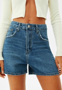 Szorty dżinsowe w średnim niebieskim kolorze z wysokim stanem, w fasonie z pięcioma kieszeniami i postrzępionymi brzegami. Materiał o fakturze z widocznymi przeszyciami.