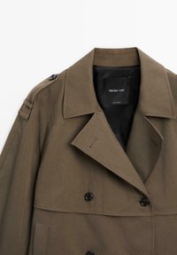 Brown trench coat featuring a double-breasted design, large lapels, stitched shoulder accents, and black buttons. Label visible inside.