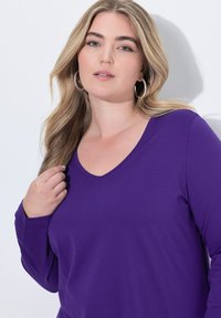 Lila Langarmshirt mit V-Ausschnitt, aus einem glatten Stoff gefertigt. Es hat lange Ärmel und eine lockere Passform.