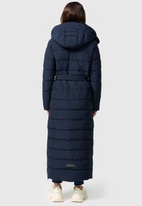 Vadderad långjacka i marinblått med huva, insvängd midja och vertikal quiltning. Har en logotyp nära fållen och rak skärning.