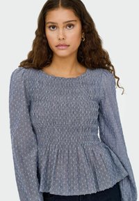Gerafftes graues Oberteil mit langen Ärmeln, strukturiertem Stoff und hellblauen Punkten. Peplum-Stil mit rundem Ausschnitt.