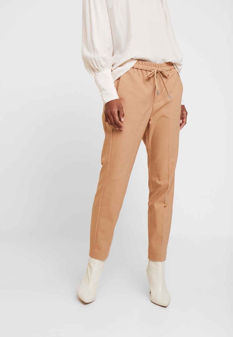 Kvinna iklädd beige dragstringsbyxor, vit långärmad blus med knappslå och vita ankelstövlar mot en neutral bakgrund.