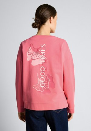 Vrouw draagt roze sweatshirt met bloemmotief en tekst "GOOD TIMES" op de achterkant, gecombineerd met donkerblauwe spijkerbroek, gezien van achteren.