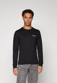 Črna majica z dolgimi rokavi iz bombaža, z okroglim izrezom in rdečim vezenim logotipom na prsih, nošena z sivimi jeans hlačami.