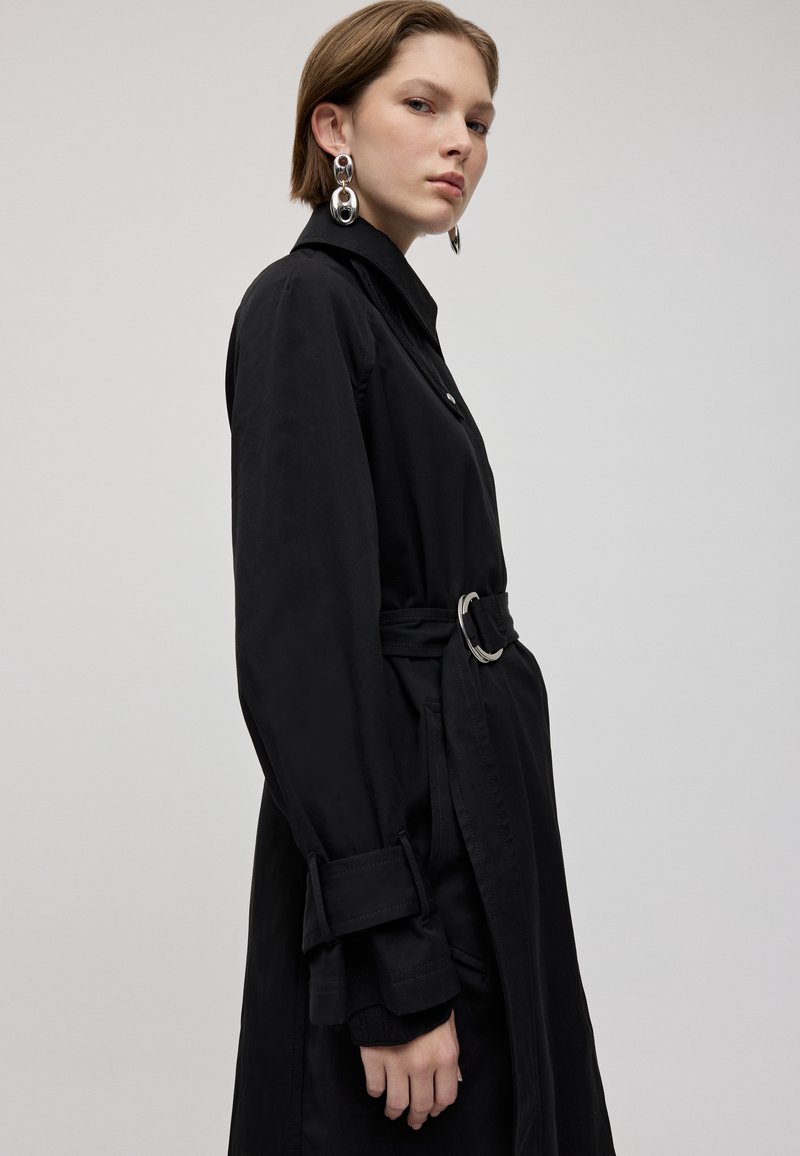 Schwarzer Trenchcoat mit Gürtel und langen Ärmel, ausgestattet mit Manschettendetails und einem silbernen Ringakzent am Gürtel, glatte Stofftextur.