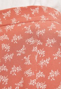 Tissu terracotta avec un imprimé floral en blanc. Présente une taille plate et une texture lisse, avec de petits motifs de feuilles et de fleurs sur l'ensemble.
