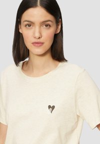 Kremowa bawełniana koszulka z dopasowanym okrągłym dekoltem i krótkimi rękawami, z małym czarnym sercem na lewej piersi.