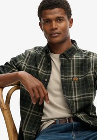 Groen en marineblauwe geruite flanellen overhemd met button-down kraag, een crèmekleurig t-shirt eronder en blauwe denim jeans, gedragen door een zittende man.
