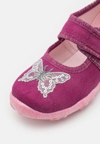 Lila tygskon med en rosa glittrande sula, prydd med en silverfärgad fjäril i paljetter. Justerbar rem för stängning.