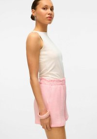 Lichte, mouwloze witte top gecombineerd met textuurvolle, roze shorts met een elastische tailleband en zijzakken. Draagt een armband.