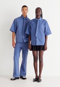 Două modele poartă ținute coordonate. Modelul masculin are o cămașă cu mânecă scurtă texturată de culoare albastră și pantaloni asortați. Modelul feminin poartă o bluză albastră și o fustă neagră.