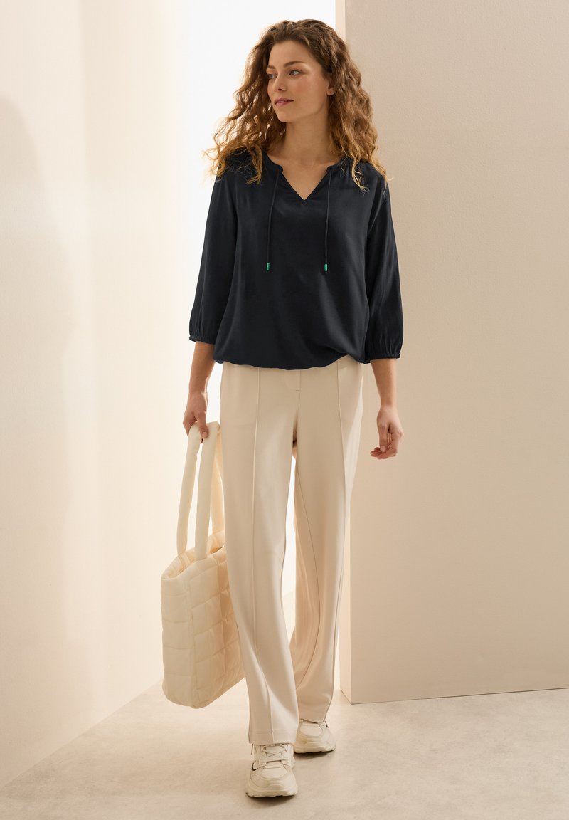 Marineblauw v-hals top met een trekkoord en puffy mouwen; gecombineerd met crèmekleurige wijde broek en een gequilt crèmekleurige handtas; witte sneakers.