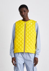 Gilet jaune matelassé avec une fermeture éclair devant, texture rembourrée, col rond et logo blanc; porté sur une chemise à manches longues bleu clair.