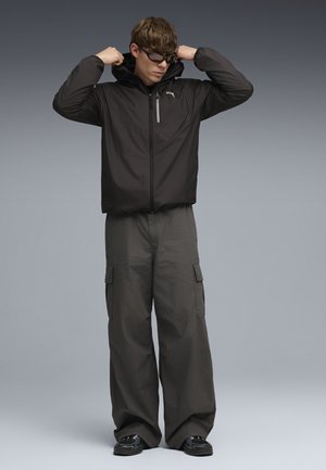 Jonge man die de capuchon van een zwarte jack met ritssluiting aanpast, draagt donkere cargobroek en zonnebril, staand tegen een grijze verloopachtergrond.