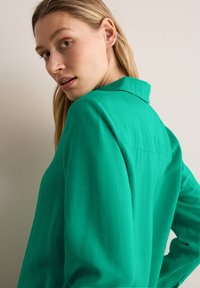 Zielona bluzka z kołnierzykiem wykonana z teksturowanej tkaniny. Posiada luźny krój, szczelinę po bokach oraz subtelną detalizację w tylnej części.
