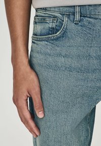 Jeans en denim bleu clair avec un gros plan sur une poche, présentant un léger délavage et une texture douce, avec une main reposant sur le tissu.