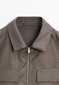 Giacca grigia di lana con colletto, chiusura frontale a zip e due tasche sul petto. Il tessuto ha una finitura testurizzata e un motivo sottile.