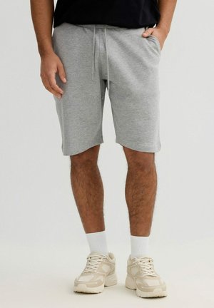 Hombre con pantalones cortos gris claro con cordón, calcetines blancos y zapatillas beige, de pie con las manos cerca de los bolsillos.