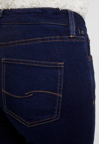 Jeans de denim azul oscuro con forma ajustada, que cuentan con un bolsillo trasero con costura curva y acentos de hilo amarillo contrastante.