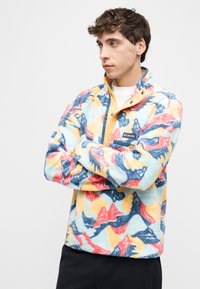 Νεαρός άνδρας με καστανά, σγουρά μαλλιά, που φοράει πολύχρωμο φλις μπουφάν με αφηρημένα μοτίβα, στέκεται με σταυρωμένα τα χέρια, κοιτώντας προς το πλάι.