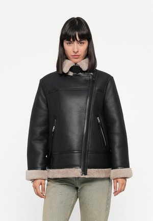 Schwarze Lederjacke mit Kunstfellkragen, Frontreißverschluss und zwei seitlichen Reißverschlusstaschen. Die Jacke hat ein weiches beiges Futter und ein kastiges Design.