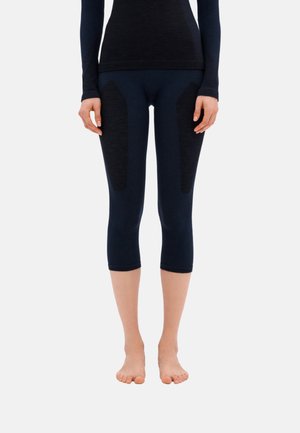 Donna che indossa leggings neri aderenti corti con pannelli testurizzati sulle cosce, in piedi a piedi nudi su uno sfondo bianco semplice.