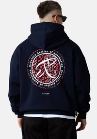 Personne portant un sweat à capuche noir avec un logo circulaire en imprimé léopard rouge et le texte blanc "Atelier créatif des Esthétiques" dans le dos, lunettes de soleil noires visibles.