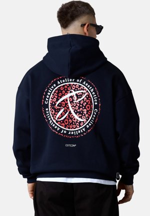 Personne portant un sweat à capuche noir avec un logo circulaire en imprimé léopard rouge et le texte blanc "Atelier créatif des Esthétiques" dans le dos, lunettes de soleil noires visibles.