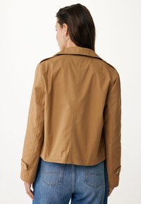 Giacca marrone corta con una texture liscia e maniche abbottonate. Include spalline e collo a scudo. Indossata sopra jeans blu.