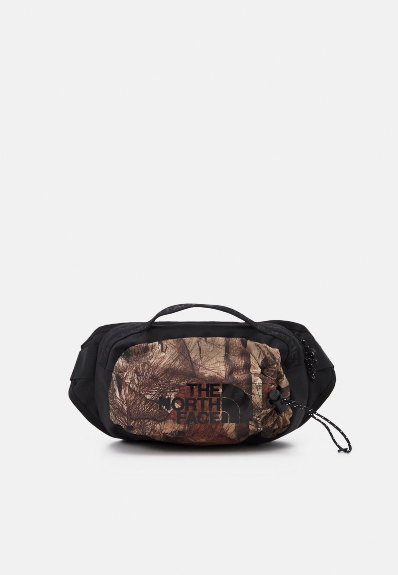Sac banane camouflage noir et marron avec poignée avant, sangle réglable et logo "The North Face" sur la poche avant.